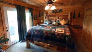 Une chambre chaleureuse aménagée dans un chalet en rondins, avec un lit en bois, une courtepointe, un ventilateur de plafond et une fresque représentant une montagne sur le mur.