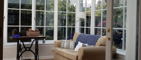 A conservatory with a beige sofa, a small table with decorative items, and a view of greenery outside.