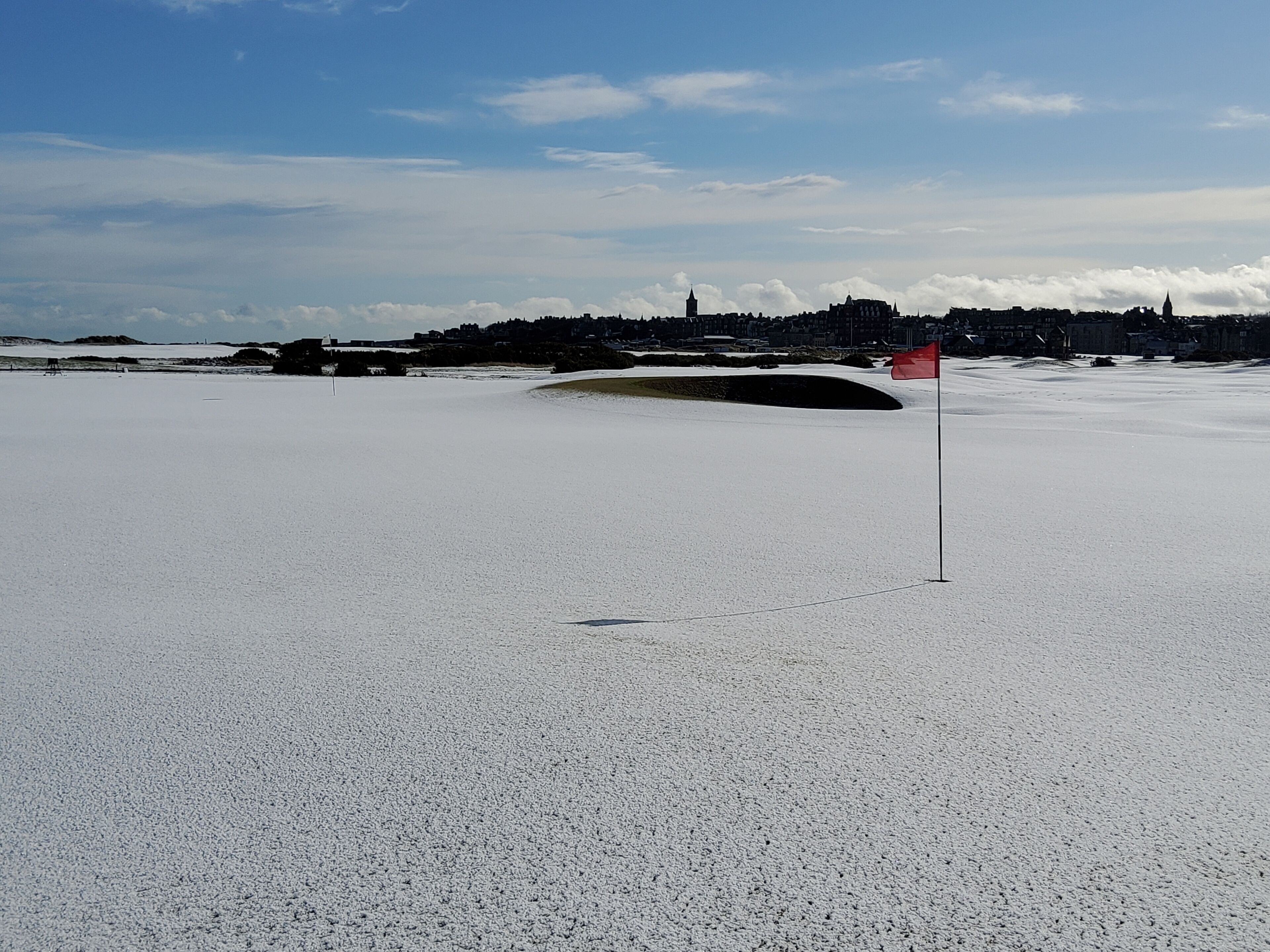 Un paysage enneigé, avec un drapeau rouge signalant un point d’intérêt, probablement un trou de golf, et un ensemble de bâtiments situé au loin.