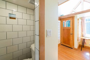 Ein Badezimmer mit gefliesten Wänden und einer Holztür, die in einen Raum mit Fenster und kleinem Tisch führt.