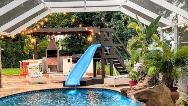 A backyard with a swimming pool, a slide, a barbecue area, and a pergola with string lights.