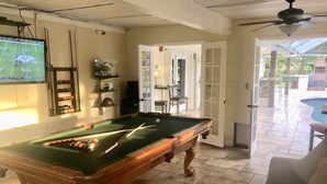 A pool table with cues and balls, a television displaying a sports game, and a door leading to an outdoor patio area with a pool.