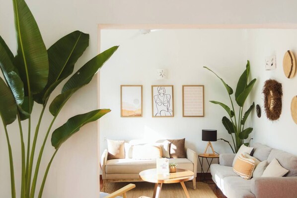 Een woonkamer met twee banken, een bijzettafeltje en grote groene planten. Er hangen kunstwerken in lijsten aan de muur en staat een lamp op een nachtkastje.