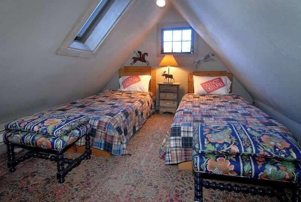 Une chambre avec deux lits, une lucarne, une petite fenêtre et une lampe.