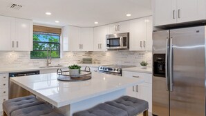 Une cuisine moderne avec des meubles blancs, des appareils en acier inoxydable, un îlot central doté de sièges et une fenêtre offrant une vue agréable.