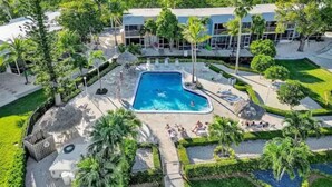Vue aérienne d’un complexe hôtelier doté d’une piscine centrale, entouré de palmiers et d’un grand bâtiment avec de nombreuses fenêtres.