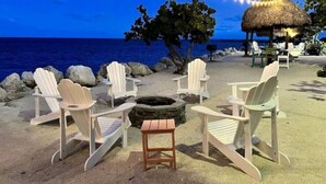 Un décor de plage avec des fauteuils Adirondack disposés autour d’un foyer, une structure à toit de chaume et des guirlandes lumineuses.