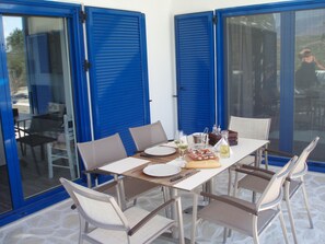 A dining table set with plates, glasses, and a bottle, surrounded by chairs, with blue shutters and glass doors in the background. - villa fully equipped with swimming pool and sea view and 360 °  (kamilari)