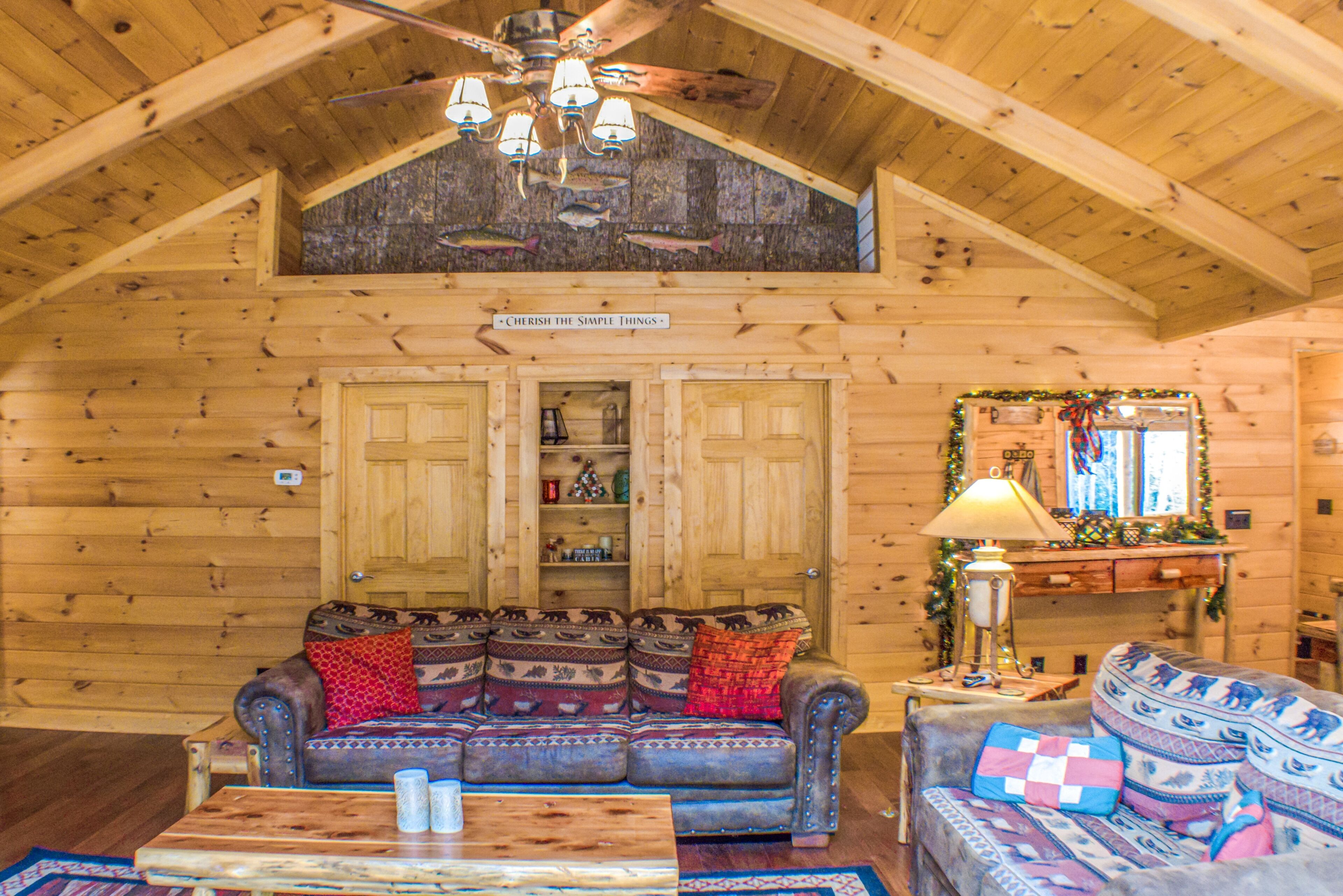 L’intérieur d’un chalet confortable, avec des murs en rondins de bois, un plafond en bois, un ventilateur de plafond, une cheminée et un canapé agrémenté de coussins à motifs.