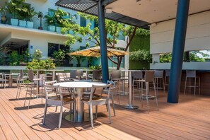 An outdoor patio area with multiple tables and chairs, surrounded by greenery and a modern building structure.