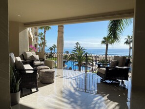Enjoy ocean views from this spacious balcony with wicker chairs and potted plants, overlooking a pool surrounded by palm trees.