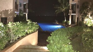 En nattbild av en swimmingpool omgiven av frodig grönska och byggnader med balkonger.