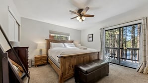 A bedroom with a wooden bed, a ceiling fan, a bench, a nightstand, and a large window with a view of trees.