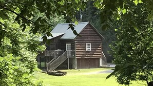 Une cabane rustique en rondins de bois, dotée d’un porche et d’un escalier menant à la porte d’entrée, entourée d’une végétation luxuriante et bordée d’un petit ruisseau.