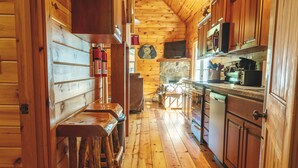 L'intérieur d'un chalet en bois comprend un espace cuisine avec des meubles en bois, une cheminée en pierre et un coin bar avec des tabourets.