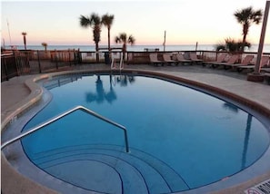 A round swimming pool with a metal ladder, surrounded by lounge chairs and palm trees, with a view of the ocean.