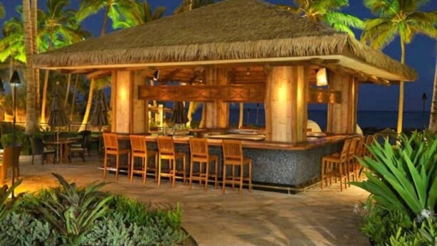 A thatched-roof bar area with wooden chairs and tables, surrounded by palm trees and a view of the ocean.