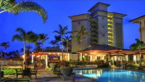 A hotel with a swimming pool, palm trees, and a well-lit building at dusk.