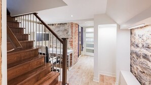 Una escalera con peldaños de madera y barandilla de metal que conduce a una habitación con pared de piedra y una puerta blanca.