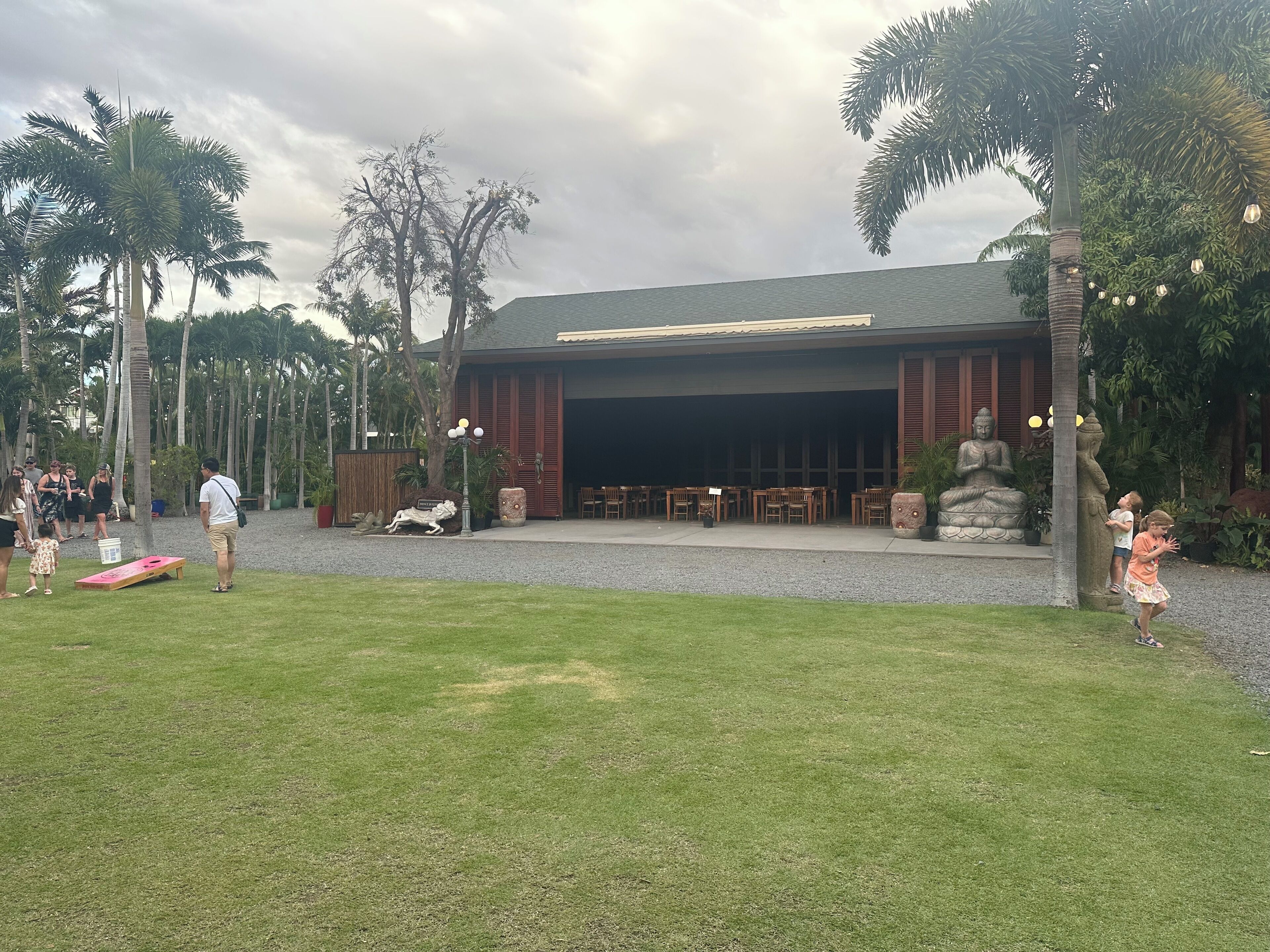 A covered outdoor area with tables and chairs, surrounded by palm trees and a Buddha statue.