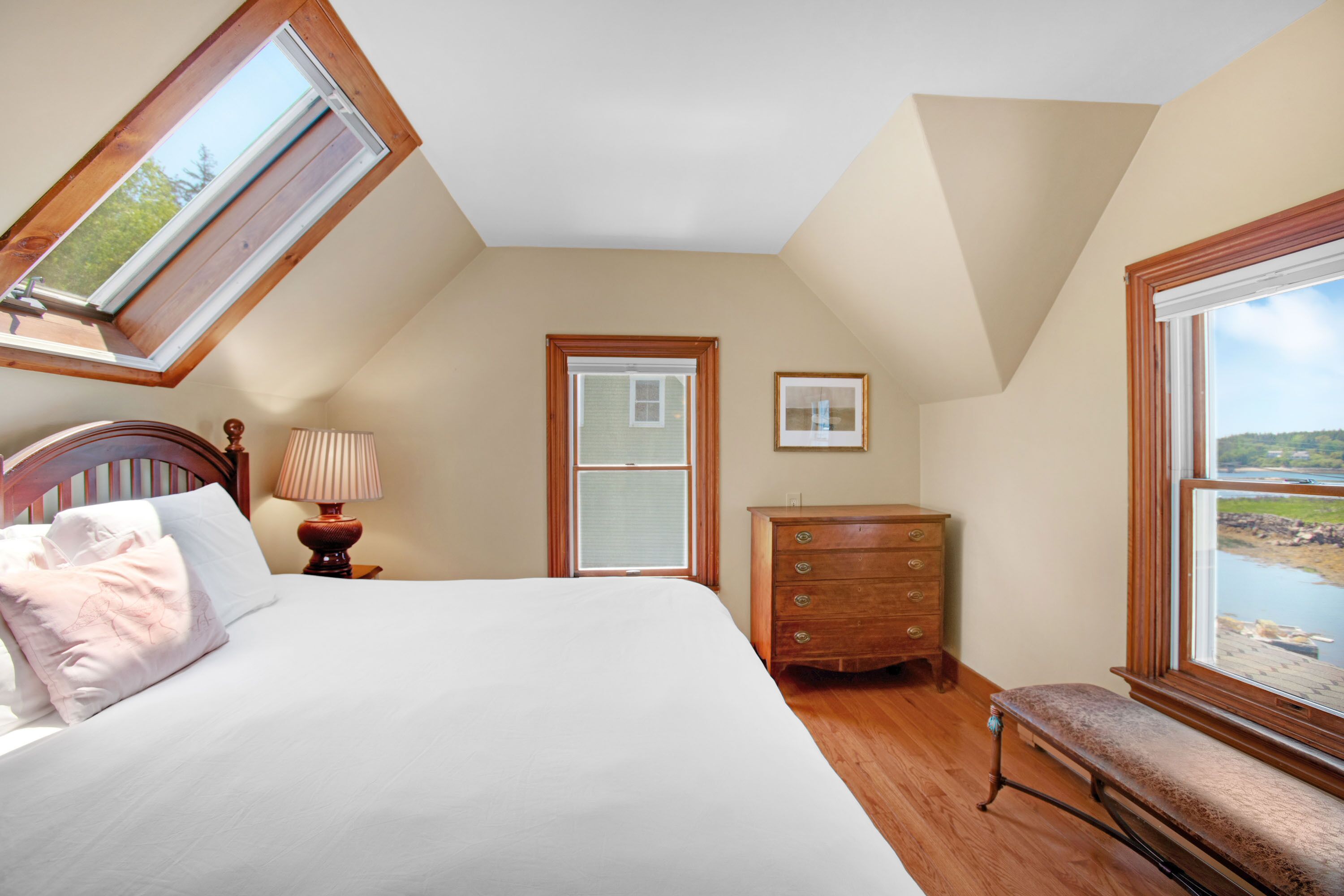 A bedroom with a skylight, a bed, a dresser, and a bench by a window overlooking a body of water.