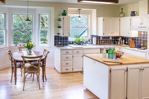 A kitchen with white cabinets, a wooden dining table, and a view of the outdoors through large windows.