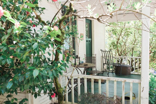 A covered porch with a table and chairs, surrounded by lush greenery and a white fence.