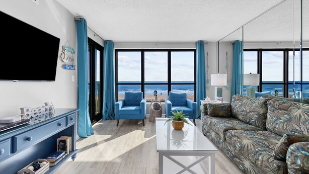Un vaste salon doté d’une grande fenêtre donnant sur la plage, d’un téléviseur à écran plat fixé au mur et d’un coin salon avec des fauteuils bleus et un canapé à motifs.