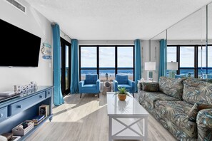 Un vaste salon doté d’une grande fenêtre donnant sur la plage, d’un téléviseur à écran plat fixé au mur et d’un coin salon avec des fauteuils bleus et un canapé à motifs.