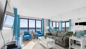 Un salon moderne doté d’une grande fenêtre donnant sur l’océan, d’un canapé bleu, d’un canapé à motifs et d’une table basse blanche.