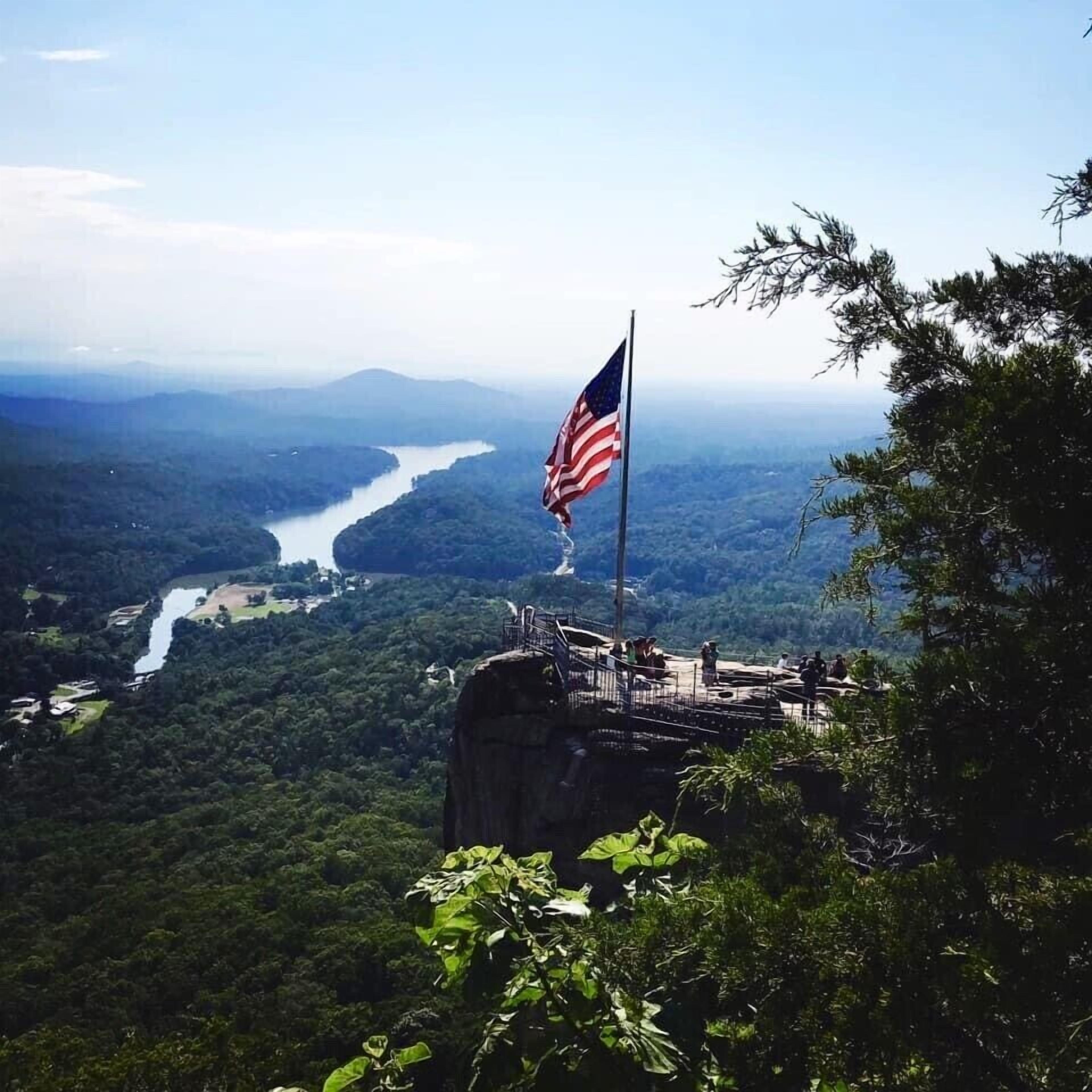 Een vlaggenmast met een Amerikaanse vlag op een rotsachtige uitkijkpost met uitzicht op een rivier en een bebost dal.