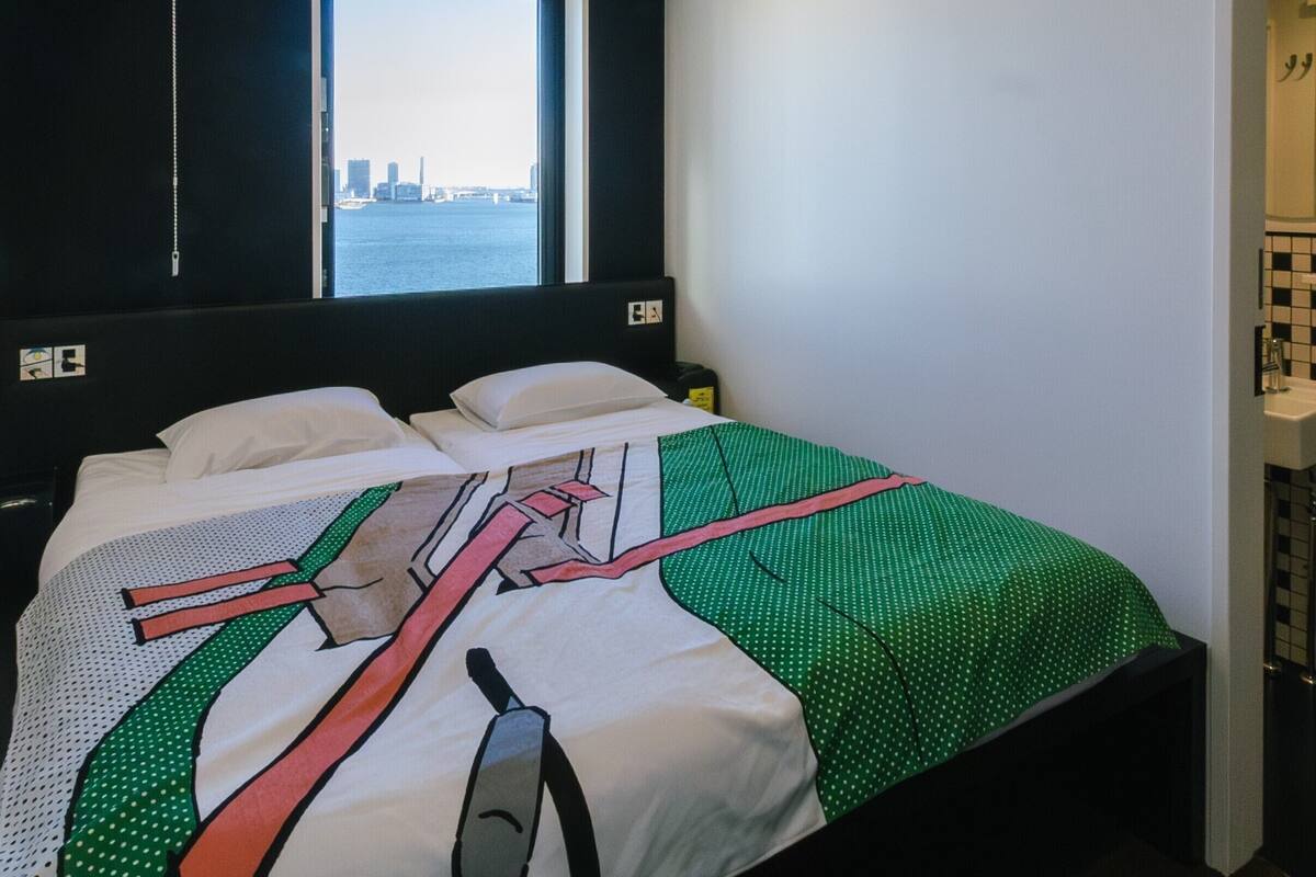 A hotel room with a bed, a window with a city view, and a bathroom visible through an open door.