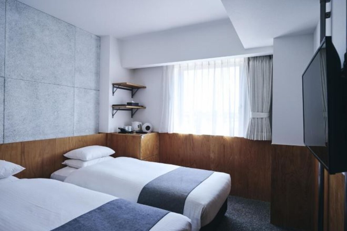 Una habitación de hotel con dos camas, televisor, ventana con cortinas y una estantería con objetos.