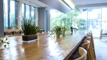 Una larga mesa de madera con sillas, plantas en macetas y vistas al exterior a través de amplias ventanas.