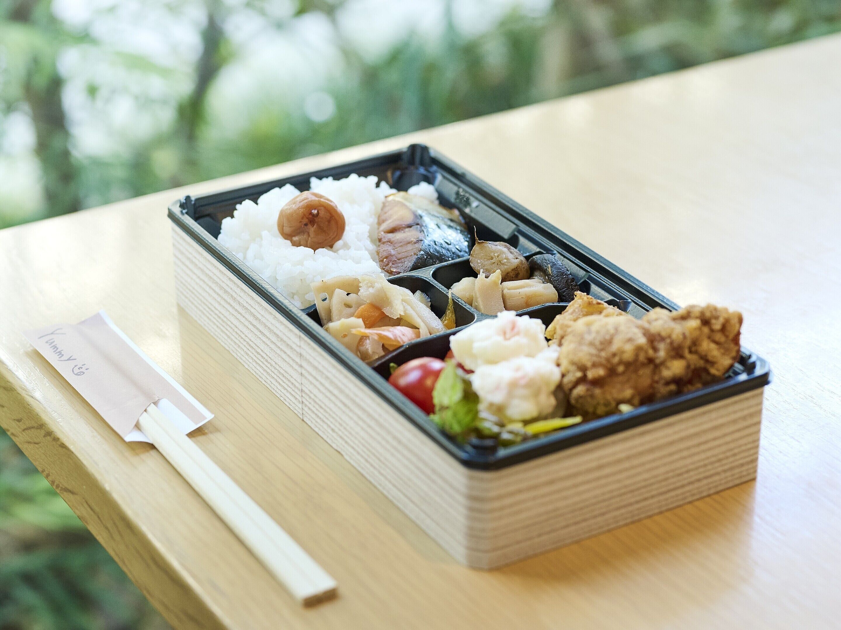 Une boîte à bento contenant du riz, du poisson, des légumes et un morceau de poulet frit, posée sur une table en bois.