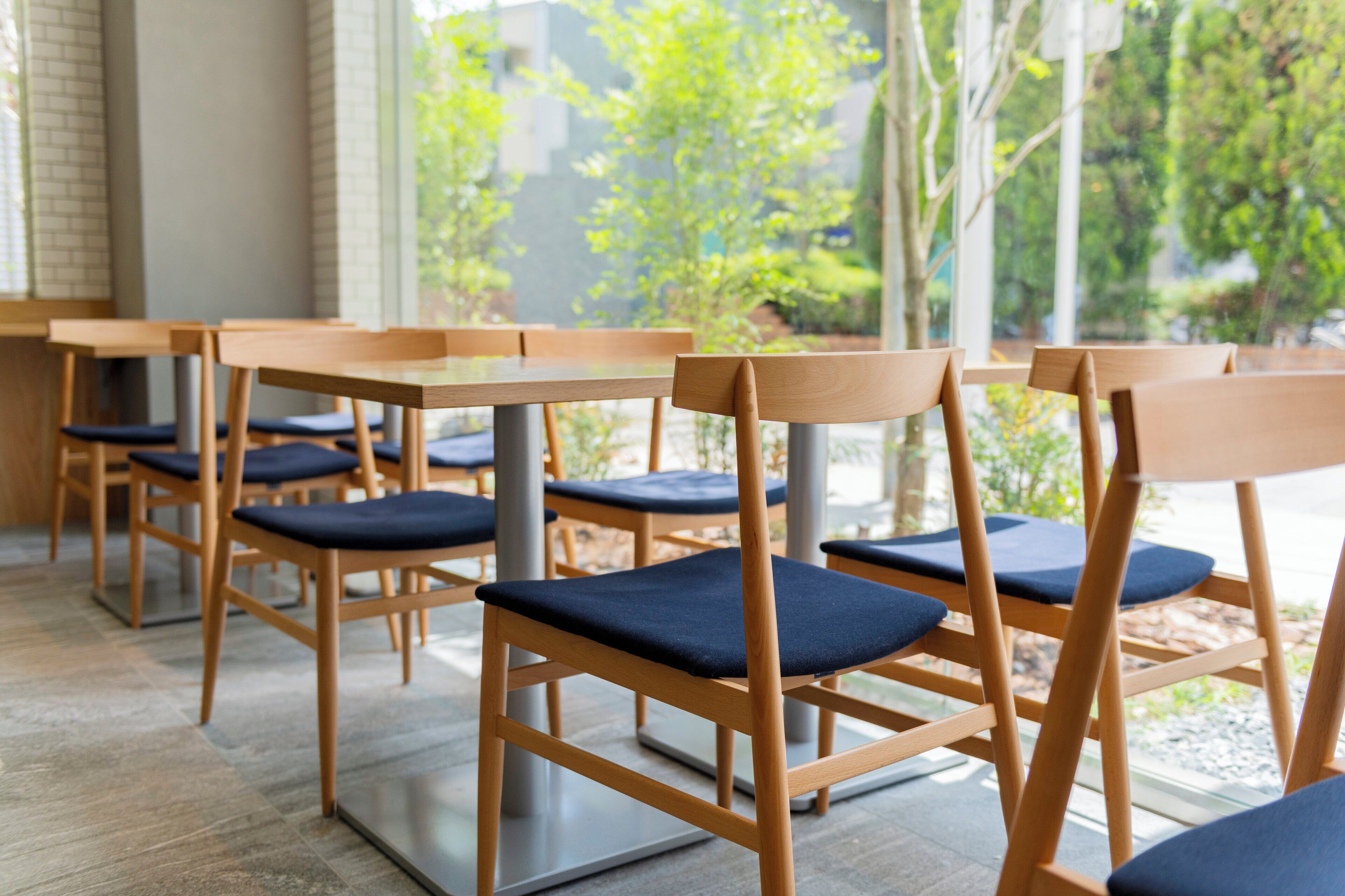 A row of wooden tables and chairs with dark blue cushions, arranged indoors with a view of greenery outside.