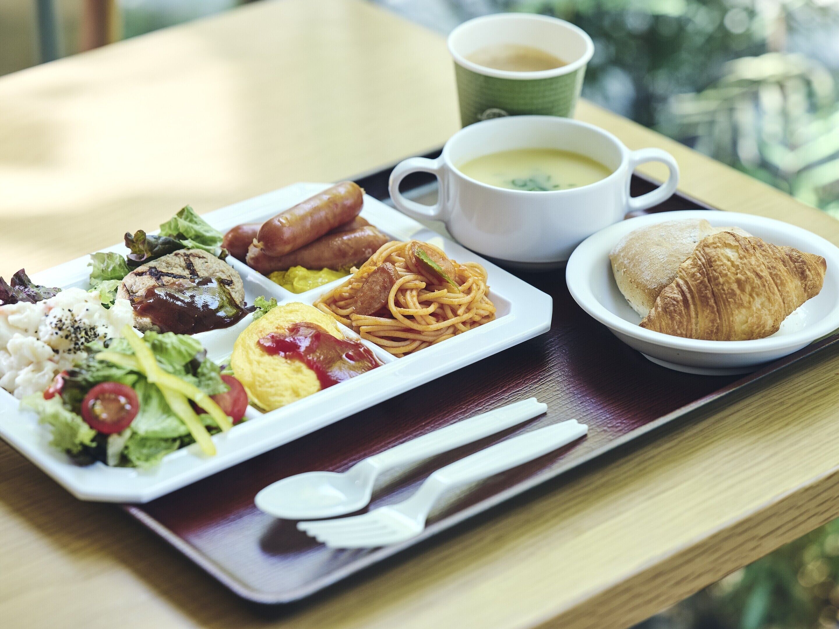 A meal tray with various dishes including pasta, sausages, a croissant, and a cup of coffee.