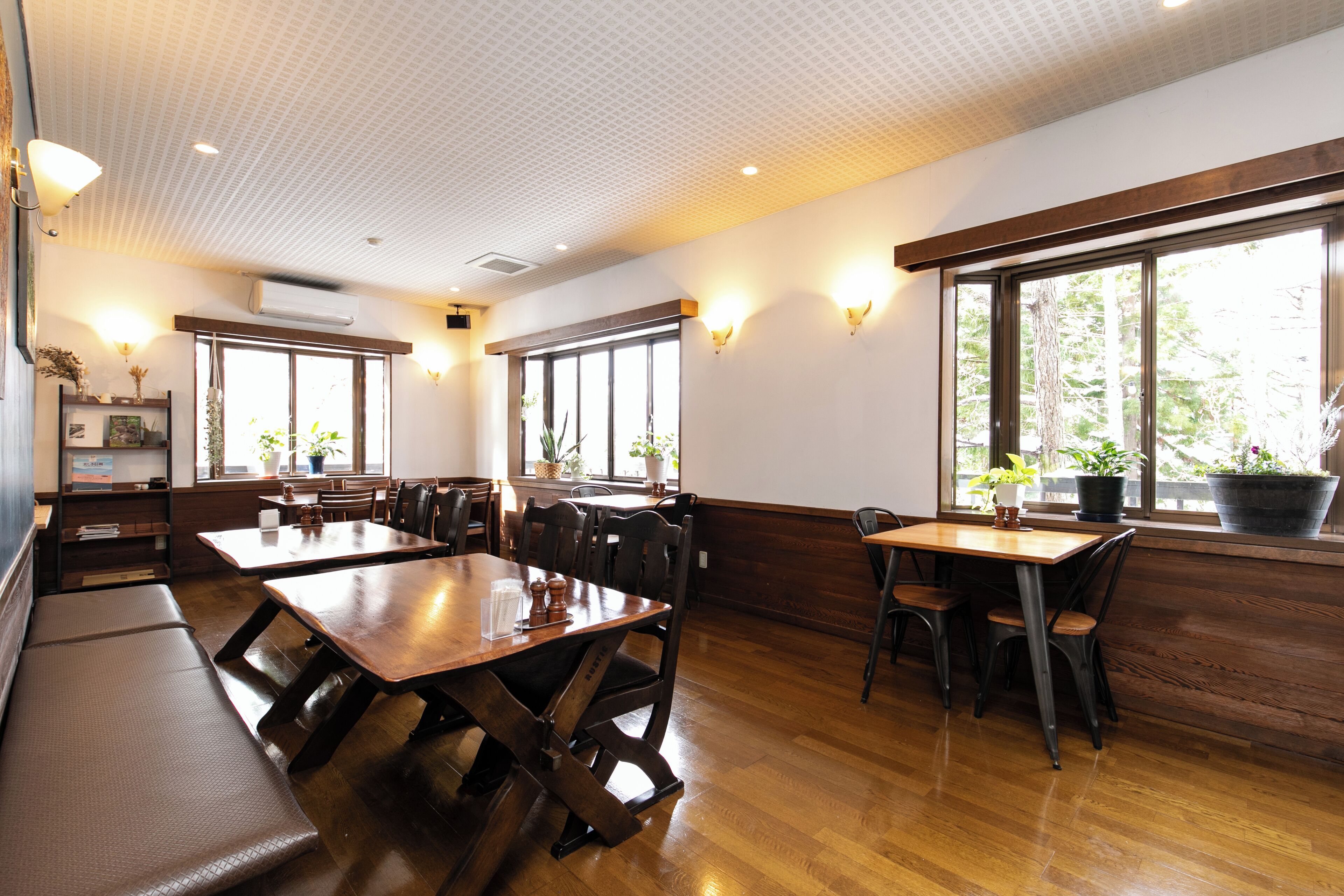 A restaurant interior with wooden tables and benches, large windows with plants, and wall-mounted lighting.