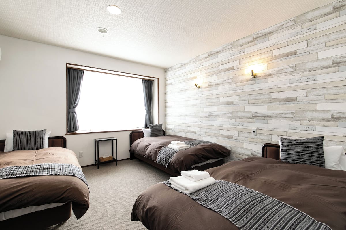 Habitació d'hotel amb dos llits, una finestra gran amb cortines, paret de relleu de maó i una taula petita.