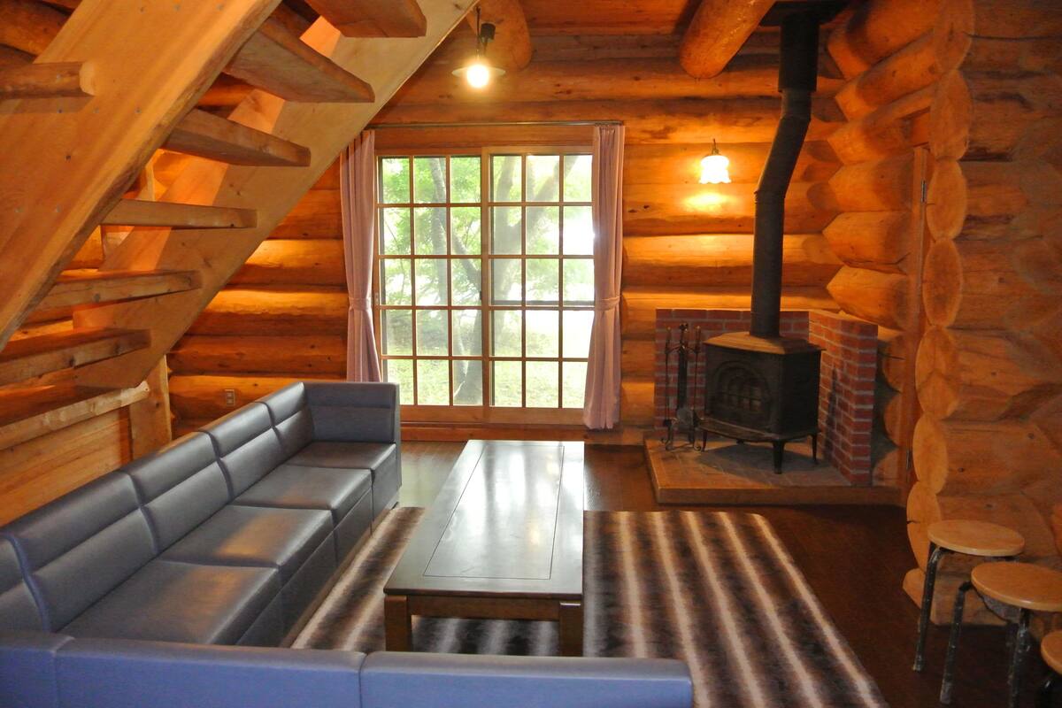 Interior d'una cabana de fusta amb un sofà, una taula, una llar de foc i una finestra gran.