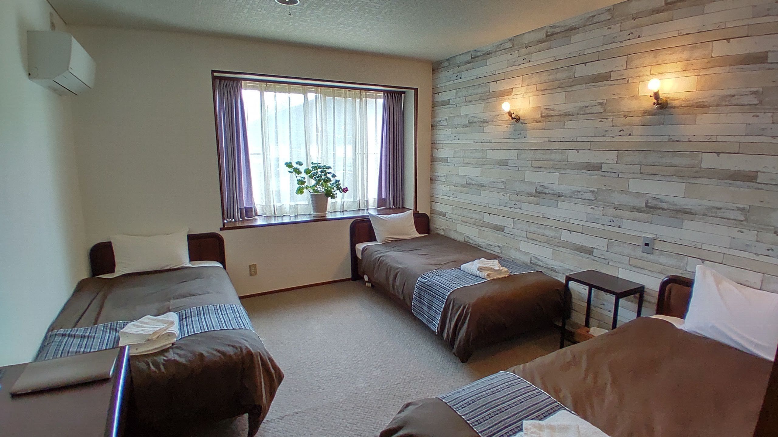 Una habitación con dos camas, una ventana con cortinas, una pared de ladrillo y una mesita.