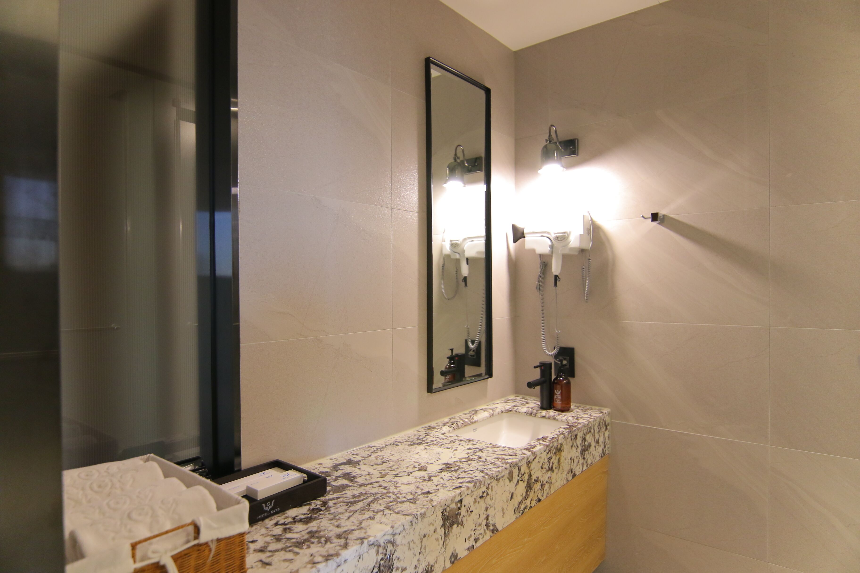 Un baño con lavamanos de mármol, un espejo, una luz empotrada en la pared y un secador de manos.