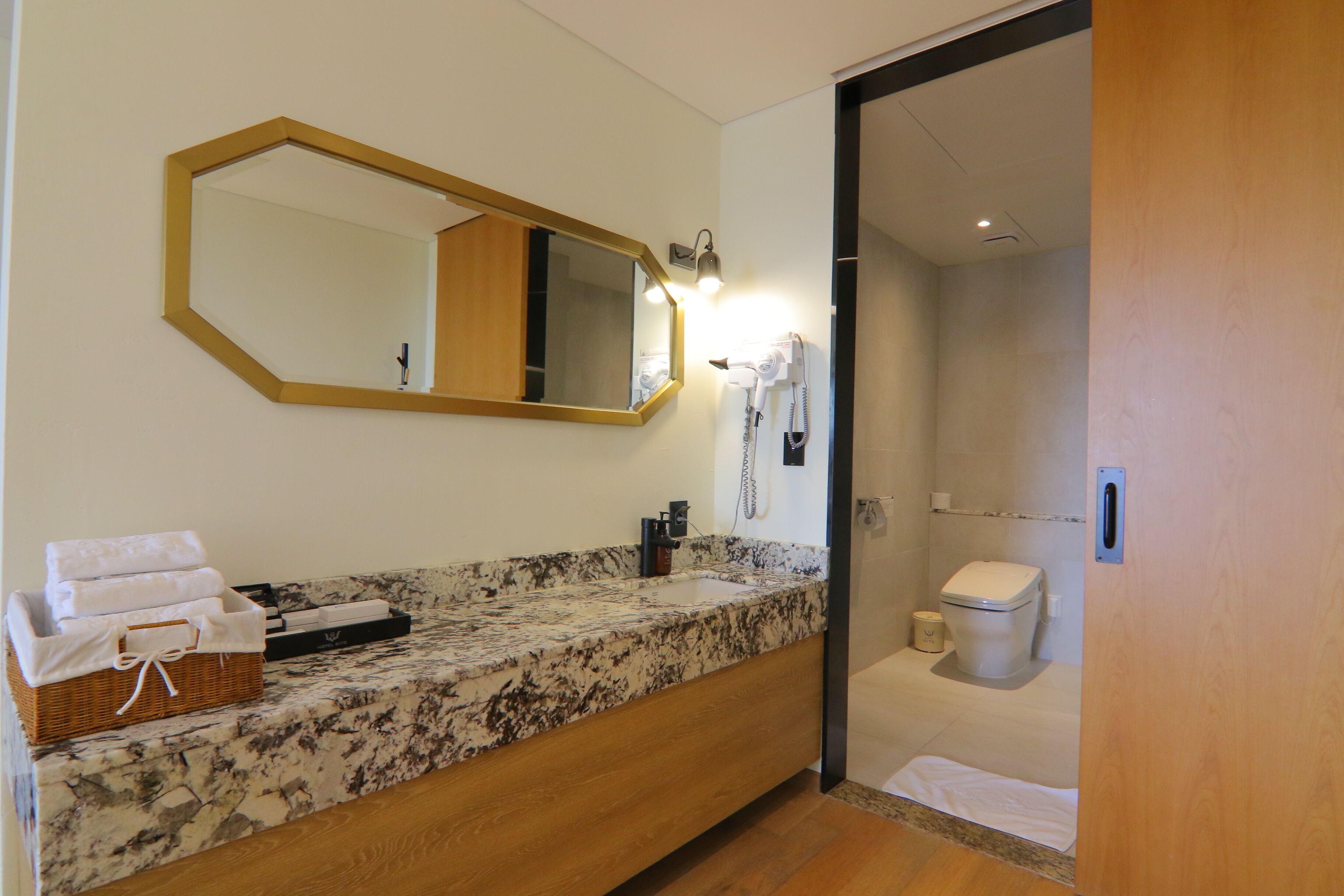 A bathroom with a marble countertop, a wooden vanity, a large mirror, a hairdryer, and a toilet.