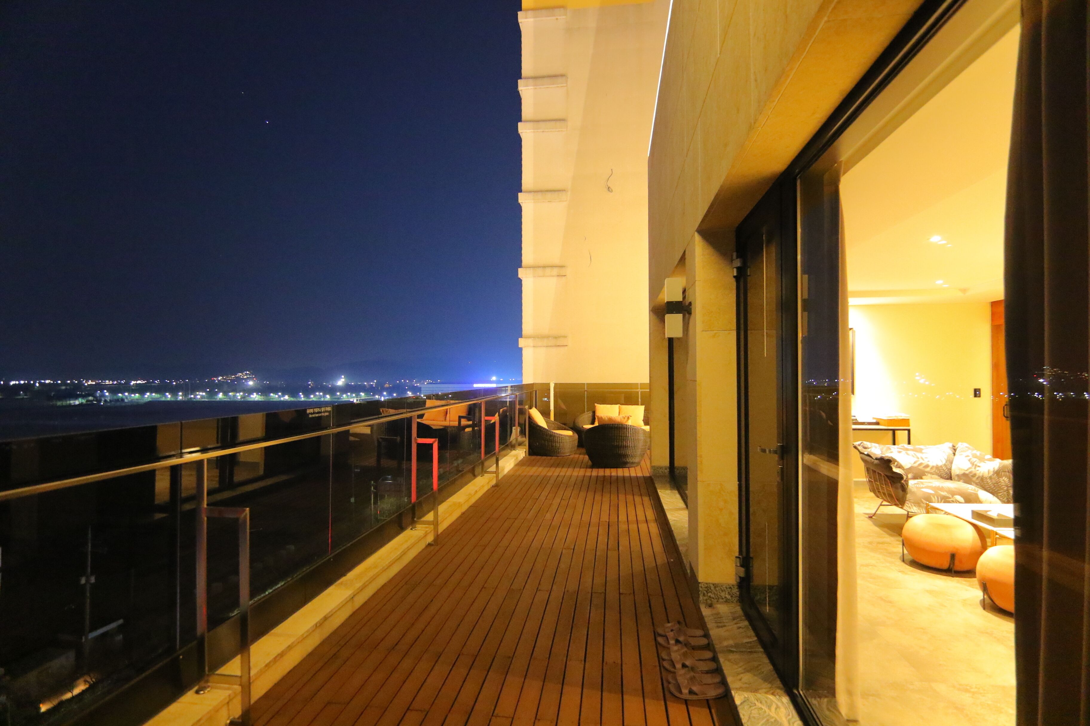 Un balcón con piso de madera, baranda de vidrio y vista de la ciudad de noche.