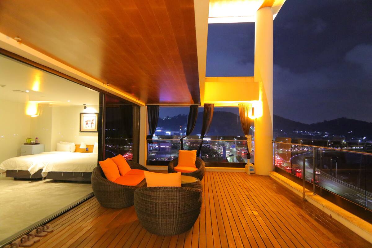 Un balcón con una cama, sillas de mimbre y vistas nocturnas de la ciudad.