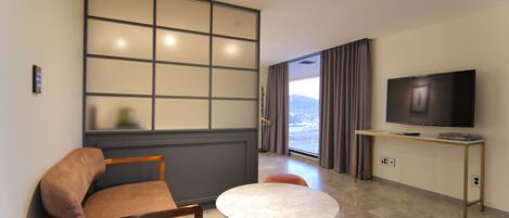 Una habitación de hotel moderna con sofá, una mesa redonda, televisor sobre un soporte y vistas al exterior a través de un ventanal.