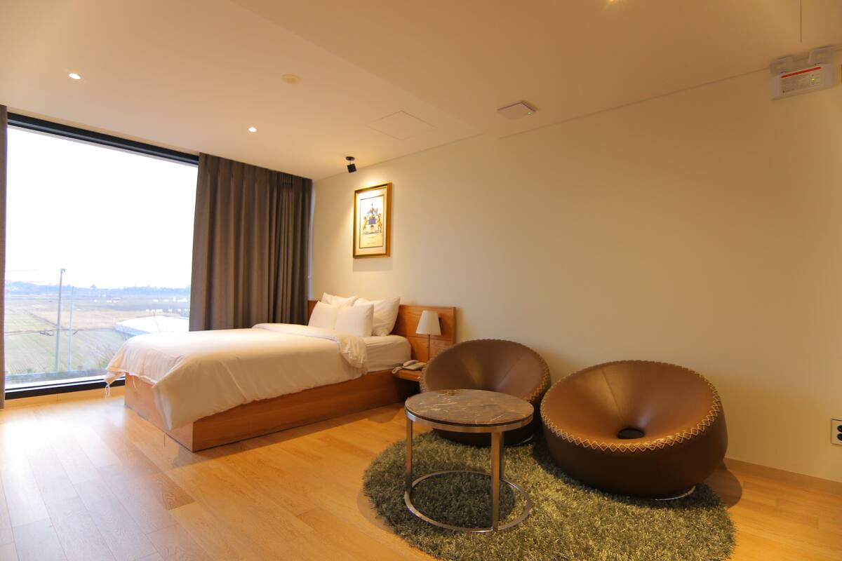 Una habitación de hotel moderna con una cama grande, una mesita redonda pequeña, dos sillas, una mesita de noche y un ventanal con vistas.