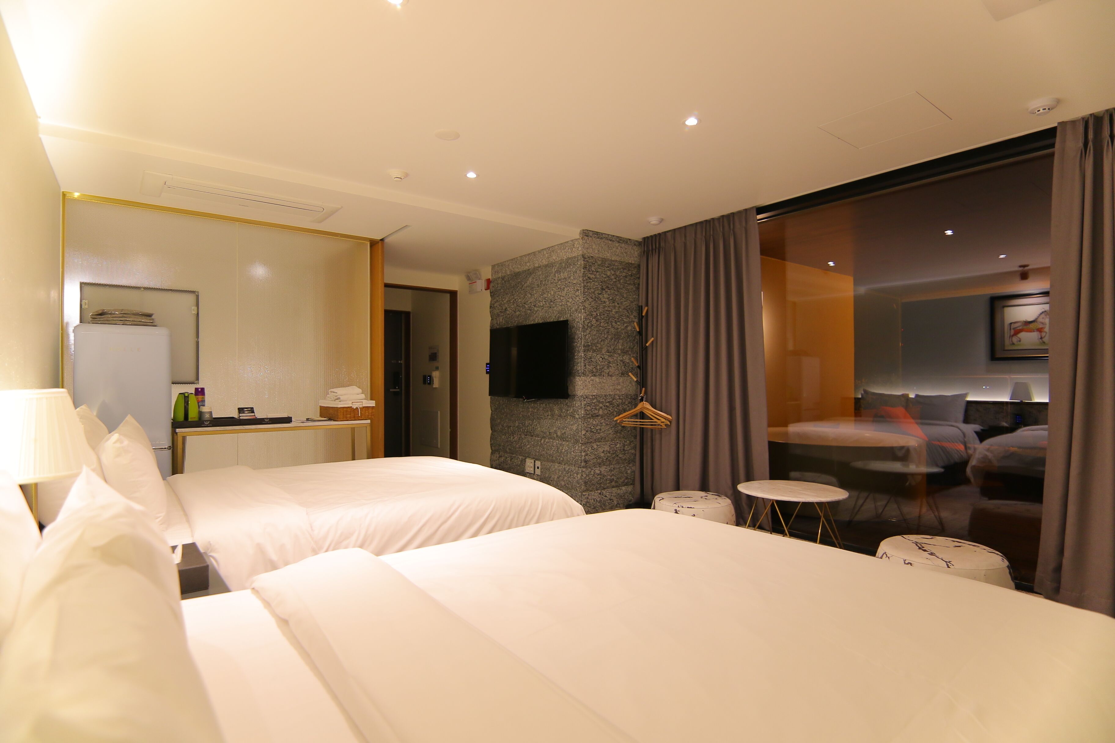 Habitación de hotel con dos camas, una mesita, televisión y un separador de vidrio con cortinas.
