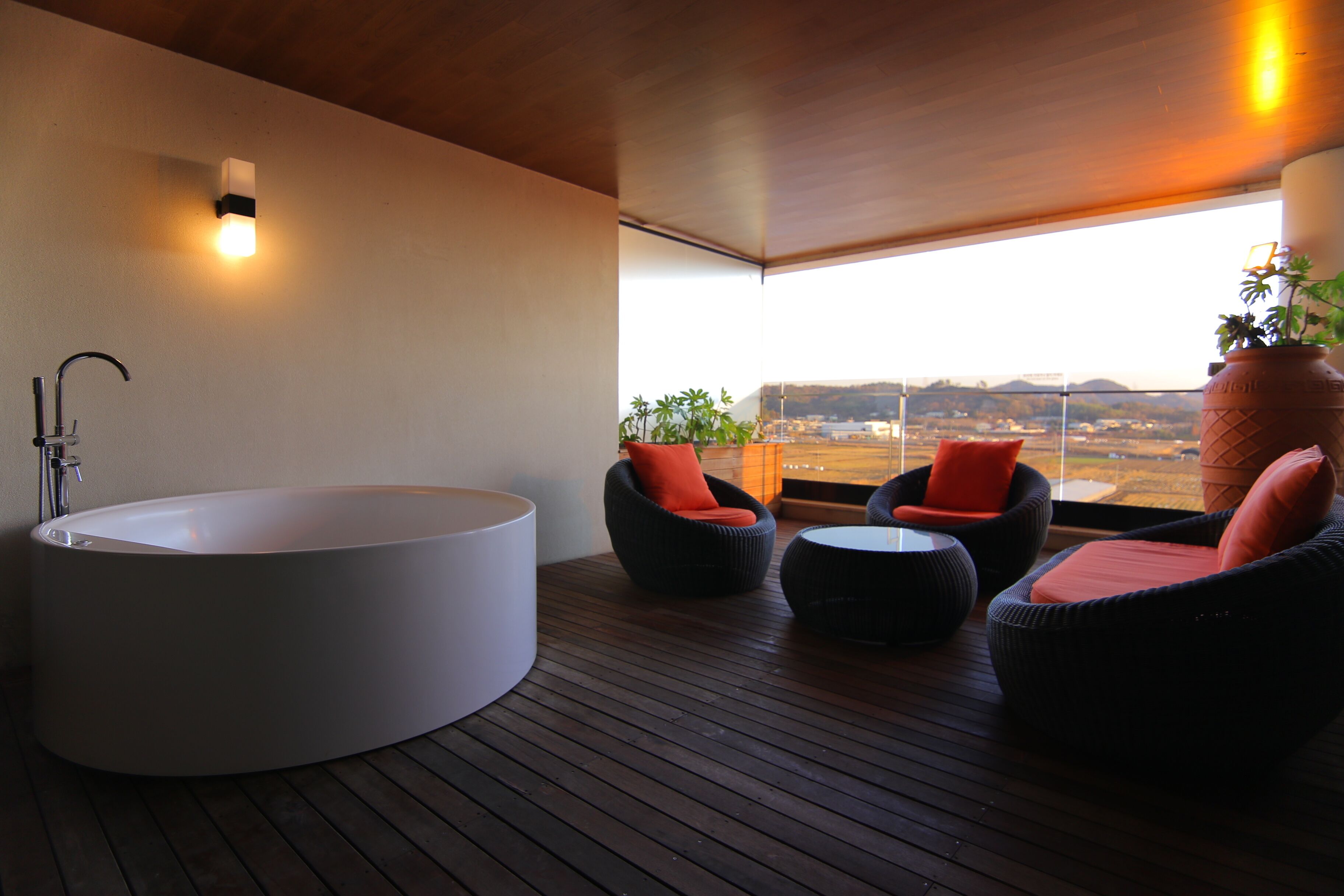A modern bathroom with a large bathtub, two wicker chairs with orange cushions, and a view of a distant landscape through large windows.