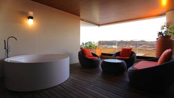 A modern bathroom with a large bathtub, two wicker chairs with orange cushions, and a view of a distant landscape through large windows.